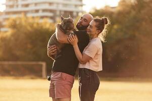 Side view, standing, and ,holding animal. Man and woman are with dog on the field photo