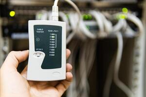 hand of system administrator or technician with tester tool for test network cables connected to patch panel of network gigabit switch in network data center. photo