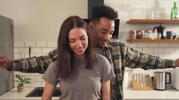 Multicultural couple in the kitchen as the camera moves from close-up to wide - woman tastes sliced tomato while man dances and hugs her from behind video