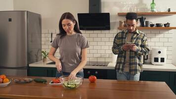 Multicultural couple dancing in the kitchen after sharing headphones - woman prepares salad while man finds music on his phone video