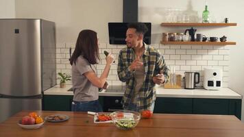 Multicultural couple singing and dancing joyfully in the kitchen - woman uses a cucumber as a microphone while man holds the phone and joins in video