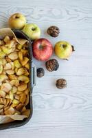 Top view of a black baking tray filled with chopped apples, fresh apples and whole walnuts on the white rustic wooden table. 03 photo