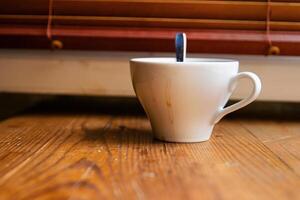 A white ceramic coffee cup with visible dried coffee stains and a metal spoon inside, placed on a rustic wooden windowsill in front of a window with closed wooden blinds. Soft focus 04 photo