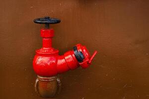 A vivid red industrial ship valve stands out against a clean brown metal wall. Big boat tap on rust painted hull surface 12 photo