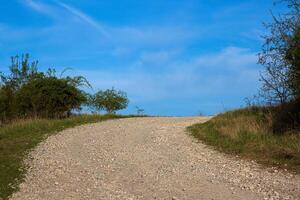 An empty gravel road winds uphill through a rural landscape. The path leads toward the horizon under a blue sky with soft clouds 18 photo