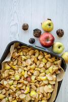 Top view of a black baking tray filled with chopped apples, fresh apples and whole walnuts on the white rustic wooden table. 04 photo