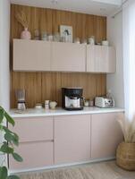 Modern kitchen with light pink and beige cabinets features coffee maker, toaster, and blender on white countertop. Decorative jars and vase add charm to wooden backsplash photo