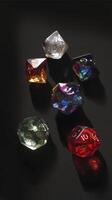 Colorful polyhedral dice arranged on dark surface, casting soft shadows. dice include various shapes and colors, such as red, blue, green, and clear, creating vibrant and playful scene photo