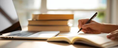 The writer's hand crafting notes by a laptop with books stacked nearby. photo