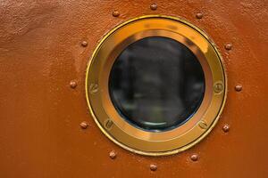 Round brass framed porthole window embedded in a textured rust colored ship wall. Contrast between polished metal and aged surface photo