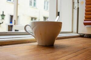 Closeup of a white ceramic coffee cup with a metal spoon, placed on a windowsill by an open window with a blurred old town of Tallinn in background. Soft focus 01 photo