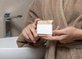 Closeup of woman in brown bathrobe with soap with a label in hands, mockup photo