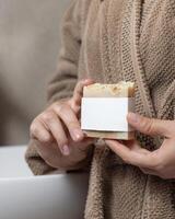 Closeup of woman in light brown bathrobe with soap bar with blank label in hands, mockup photo