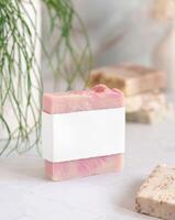 Soap bar with blank label on marble countertop near green plant close up, cosmetic mockup photo