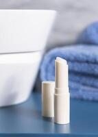 Opened lip balm stick with a lid on blue conutertop near cosmetics and basin in bathroom, mockup photo