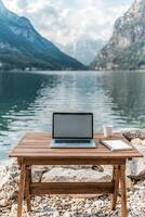 Desk with a laptop by a serene lake and mountains in the background. Work-life balance strategies concept. photo