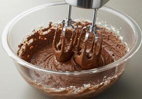 A close up of a clear bowl with chocolate batter being mixed with an electric mixer. photo