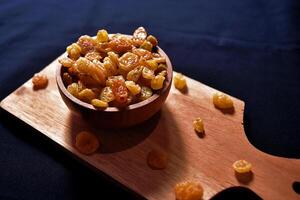 Closeup view of a bowl of raisins on a wooden cutting board with a dark background. Food photography of a bunch of dried grapes commonly used in cooking and baking. photo