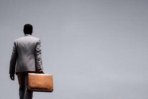 A businessman dressed in a light gray suit walks away, holding a brown briefcase. The background is smooth and neutral, emphasizing the subjects solitary journey in a corporate environment. photo