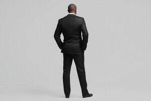 A man wearing a sleek black suit stands with his back to the viewer, exuding confidence and professionalism. The backdrop is minimalist, enhancing the focus on his attire and posture. photo
