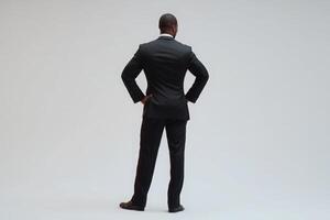 A man wearing a formal black suit stands with his back to the viewer, hands resting on hips. His posture conveys confidence. The setting features a simple and neutral background. photo