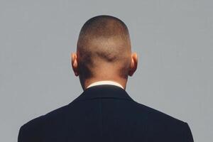 A man is seen from behind, showcasing a closely shaved head and wearing a formal suit. The focus is on his back and the simplicity of the backdrop, highlighting his posture. photo