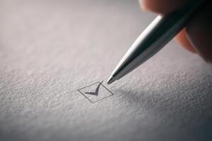 A persons hand grips a silver pen, making a precise checkmark inside a small empty box on white paper, signifying task completion during a planning or organizing session. photo
