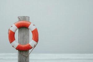 A bright orange and white lifebuoy hangs on a weathered wooden post by the ocean. The calm waters reflect the gray sky, creating a serene atmosphere on a cloudy day. photo