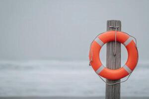 A bright orange lifebuoy hangs on a weathered wooden post at the edge of a tranquil beach. The ocean waves rest gently in the background, under a cloudy sky, creating a serene atmosphere. photo