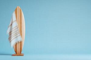 A wooden surfboard stands upright on a simple base, draped with a softly colored towel. The smooth blue background creates a calming atmosphere, reminiscent of beach vacations and sunny days. photo