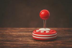 A vintage red and white joystick controller rests on an aged wooden surface. Soft lighting enhances its nostalgic appeal, evoking memories of classic gaming from the past. photo
