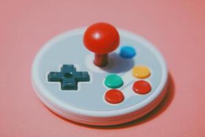 A vintage-style gaming joystick rests on a soft pink background. Bright buttons in various colors contrast with the muted design, evoking feelings of nostalgia and retro gaming experiences. photo