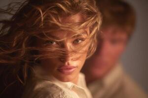 A woman with long, wavy hair stands outside, her face illuminated by soft light, as her hair is caught by the wind. A blurred figure appears in the background, creating a sense of depth and intimacy. photo