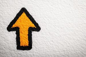 A bold orange arrow outlined in black is depicted on a textured white surface, effectively directing attention upwards. The vibrant colors contrast sharply, providing a clear visual cue. photo