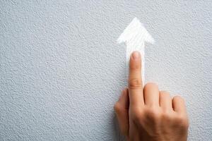 A hand with a finger extended points towards a white arrow sketch on a gray wall. The gesture suggests direction or an upward movement, creating an engaging visual focus. photo