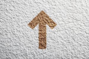 A tan arrow points upward. The arrow and background surface have a rough, textured appearance. The arrows color contrasts with the white, uneven surface. photo
