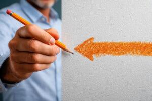 A hand holds a yellow pencil, illustrating the act of drawing an orange arrow on a textured white surface. The environment appears well-lit, focusing on the creative process. photo