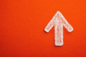 An arrow shaped figure made of white textured material is prominently displayed against a bright orange background. The contrasting colors create a striking visual emphasis on direction. photo