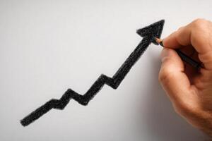 A hand holds a black pencil, sketching a jagged upward trend line on a blank white surface. This represents growth or increasing success, typically associated with financial or business progress. photo