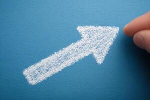 A hand is about to complete the drawing of a white arrow pointing upwards on a vibrant blue surface. The arrow symbolizes growth, progress, and direction, conveying a positive message. photo