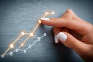 A hand with neatly manicured white nails is gently touching a glowing line graph on a textured gray background. The graph shows upward trends, symbolizing positive growth and data analysis. photo