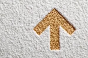 A textured arrow symbol made from golden material is prominently displayed on a white background. The design highlights the upward movement, conveying a sense of direction and progress. photo