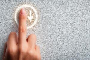 A hand gently presses a circular button on a textured grey wall. The button has a downward arrow symbol, suggesting a function related to moving or navigating downward. photo