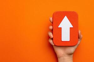 A hand grasps a rectangular orange sign featuring a large white arrow pointing upwards against a vivid orange backdrop. The composition emphasizes clarity and simplicity. photo