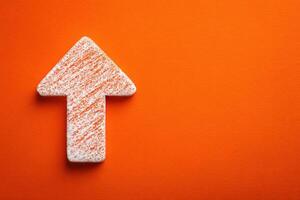 A textured arrow made of white material with orange streaks is positioned on a vibrant orange surface. The arrow points upward, symbolizing direction and growth in a minimalist setting. photo