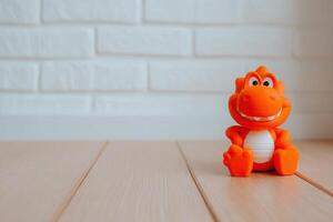 A bright orange dinosaur toy with a friendly smile is positioned on a wooden floor. The background features a light-colored brick wall, creating a simple and inviting atmosphere. photo