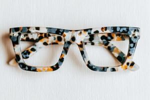 A pair of tortoiseshell glasses lays flat against a white surface. The multi-colored frames feature a distinctive pattern of brown, black, and cream, emphasizing a modern and trendy look. photo