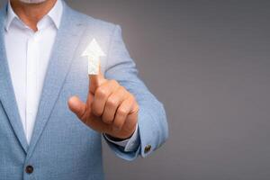 A man in a light blue suit points towards a digital upward arrow symbol, representing growth or advancement in a professional context. The backdrop is a simple gray hue. photo