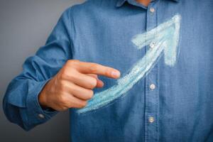 A person in a blue shirt is pointing with their finger at an upward arrow drawn in chalk on a grey wall. The gesture signifies growth and aspiration, capturing a moment of motivation and positivity. photo