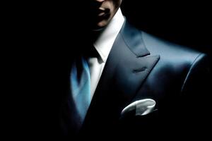 A well-dressed man in a dark suit showcases sophistication. His blue tie and white pocket square add elegance. The low-key lighting highlights his attire while obscuring his face, creating intrigue. photo